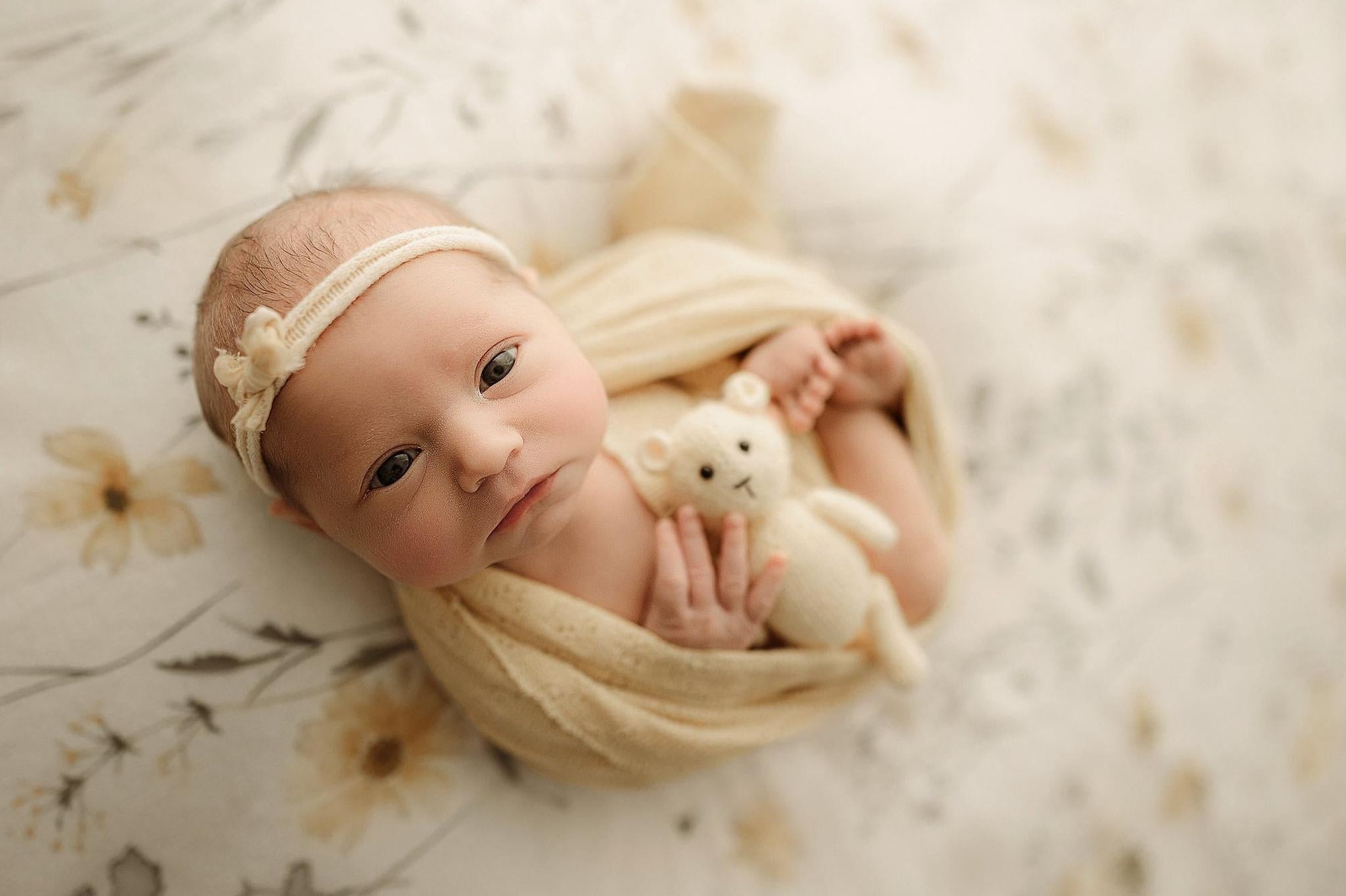 Newborn wrapped in a butter yellow knit blanket on an ivory floral fabric backdrop with a matching teddy bear.