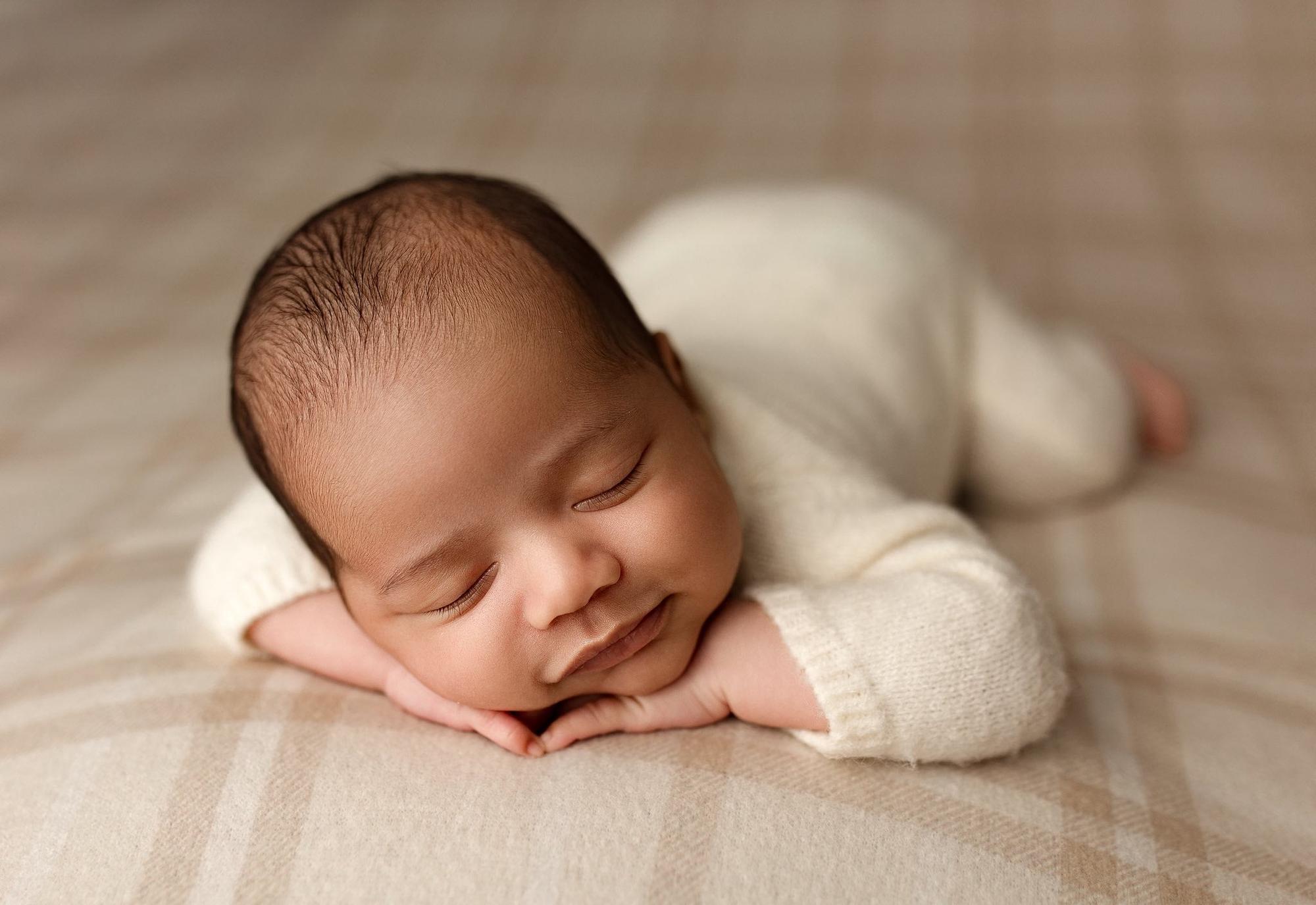 Newborn sleeping on a soft, neutral beige plaid posing fabric, wearing a cozy cream-colored knit outfit.