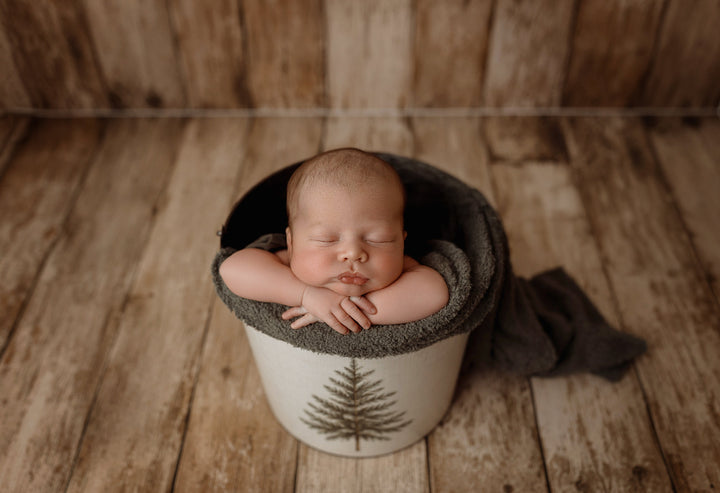 Sleeping newborn baby posed in a rustic metal bucket featuring the Little Evergreen Christmas bucket cover with a vintage pine tree design, styled with cozy dark green knit wraps and a wooden backdrop for newborn photography.