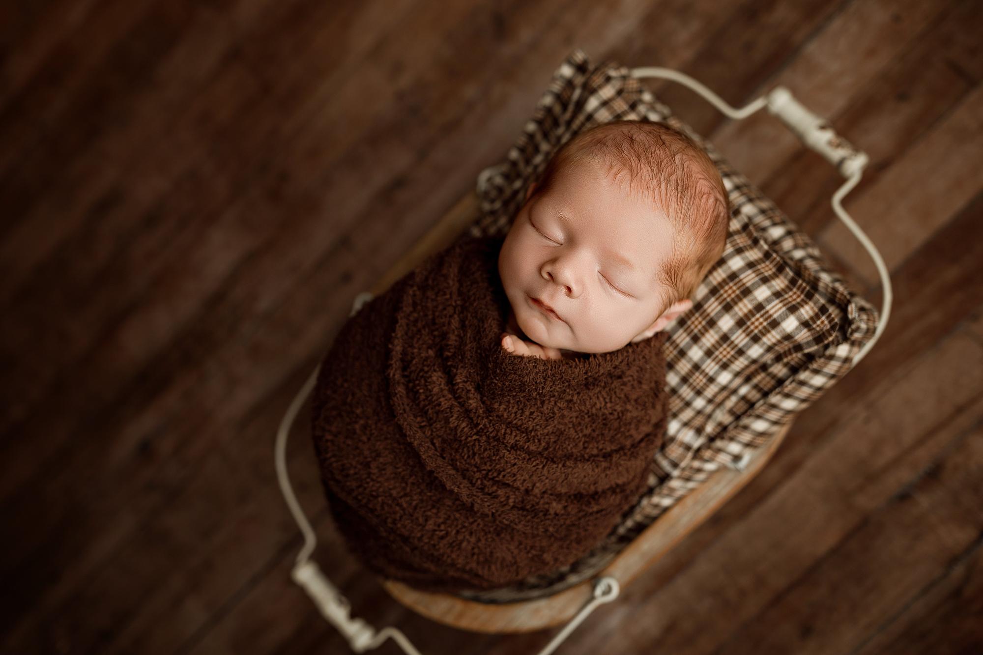 Newborn swaddled in a soft, fuzzy cocoa brown wrap, resting in a rustic basket with a plaid cushion.