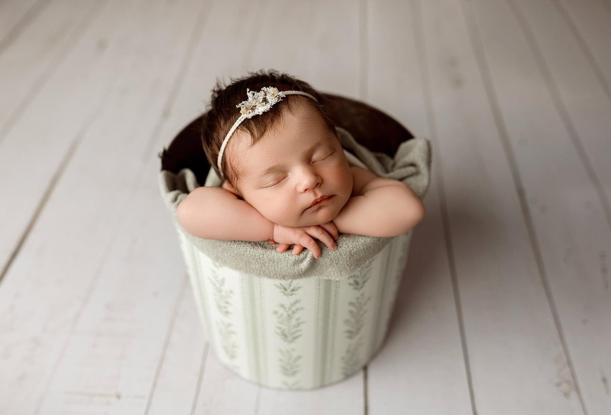 Newborn baby sleeping in a bucket wrapped in a textured, sage gray Nest fabric with organic, earthy styling.