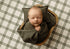 Sleeping newborn baby holding a Mantle Garland olive green mini blankie in a wooden bowl, styled with coordinating green pillows and a black and white plaid backdrop for newborn photography.