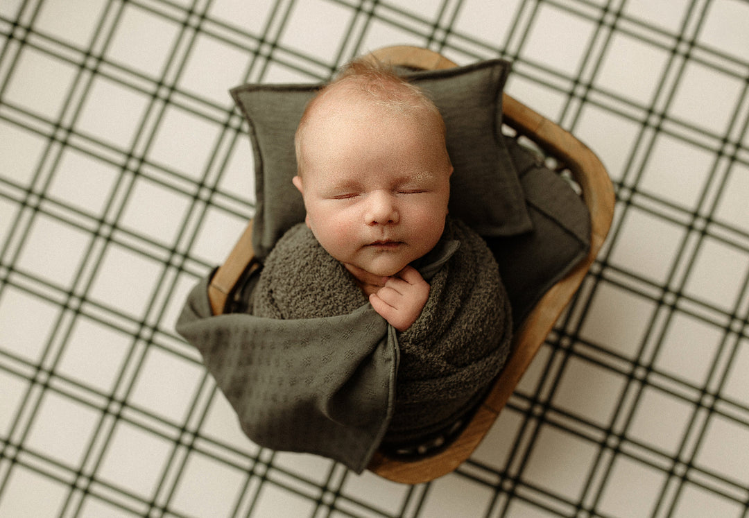 Sleeping newborn baby holding a Mantle Garland olive green mini blankie in a wooden bowl, styled with coordinating green pillows and a black and white plaid backdrop for newborn photography.