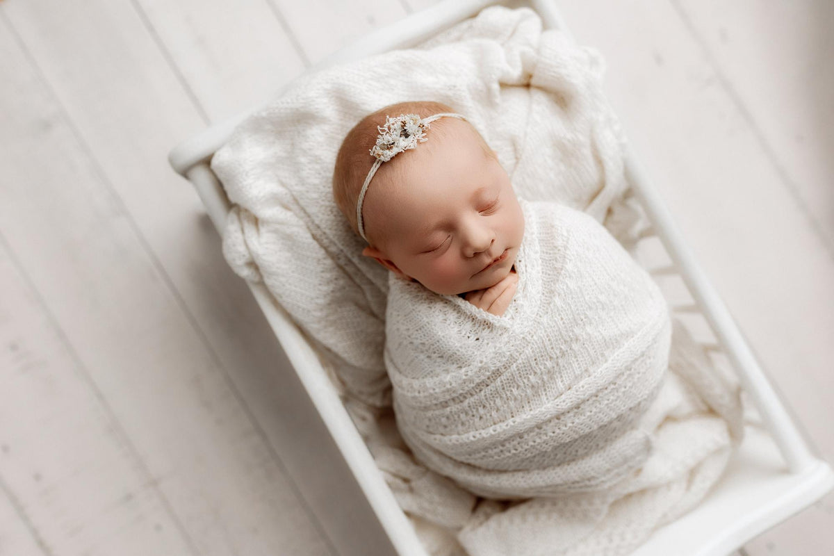 Newborn baby swaddled in soft, ivory open-knit fabric, resting in a white wooden prop bed.