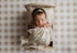 Newborn wrapped in a soft brown-purple stretch knit fabric, tucked into a wooden bed with floral ruffle bedding.