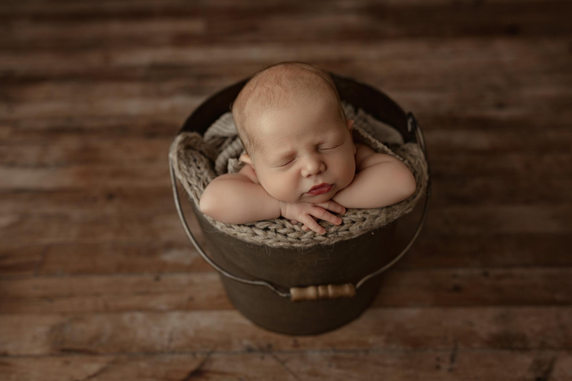 A sleeping newborn baby is posed inside a rustic dark metal bucket lined with a chunky knit blanket, set on a weathered wooden plank floor.