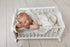 Newborn sleeping in a white wooden bed, wrapped in a soft, ivory open-knit fabric posing wrap.