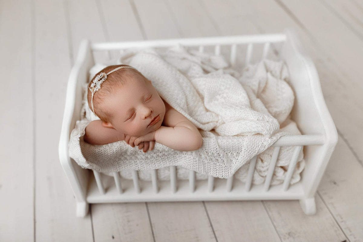 Newborn sleeping in a white wooden bed, wrapped in a soft, ivory open-knit fabric posing wrap.