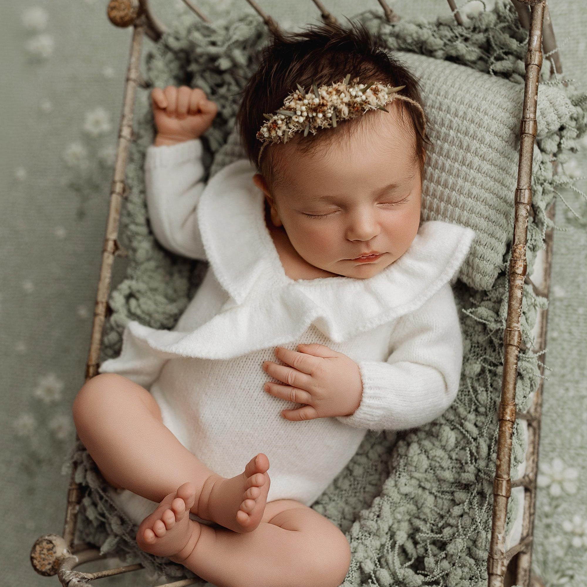 A sleeping newborn baby wearing a white ruffled long-sleeve bodysuit and a delicate floral headband, lying in a rustic twig bassinet on a textured sage green blanket with a matching patterned backdrop.