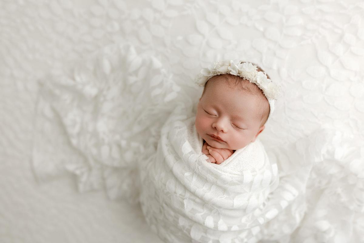 Sleeping newborn baby wrapped in ivory fabric with Minimalist lace layer and coordinating delicate ivory floral headband.