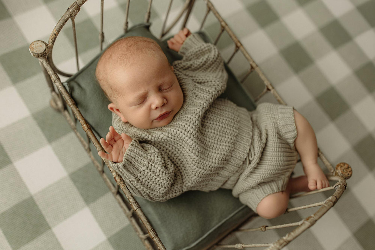 Newborn sleeping on a soft moss green velvet pillow inside a vintage metal bed frame.