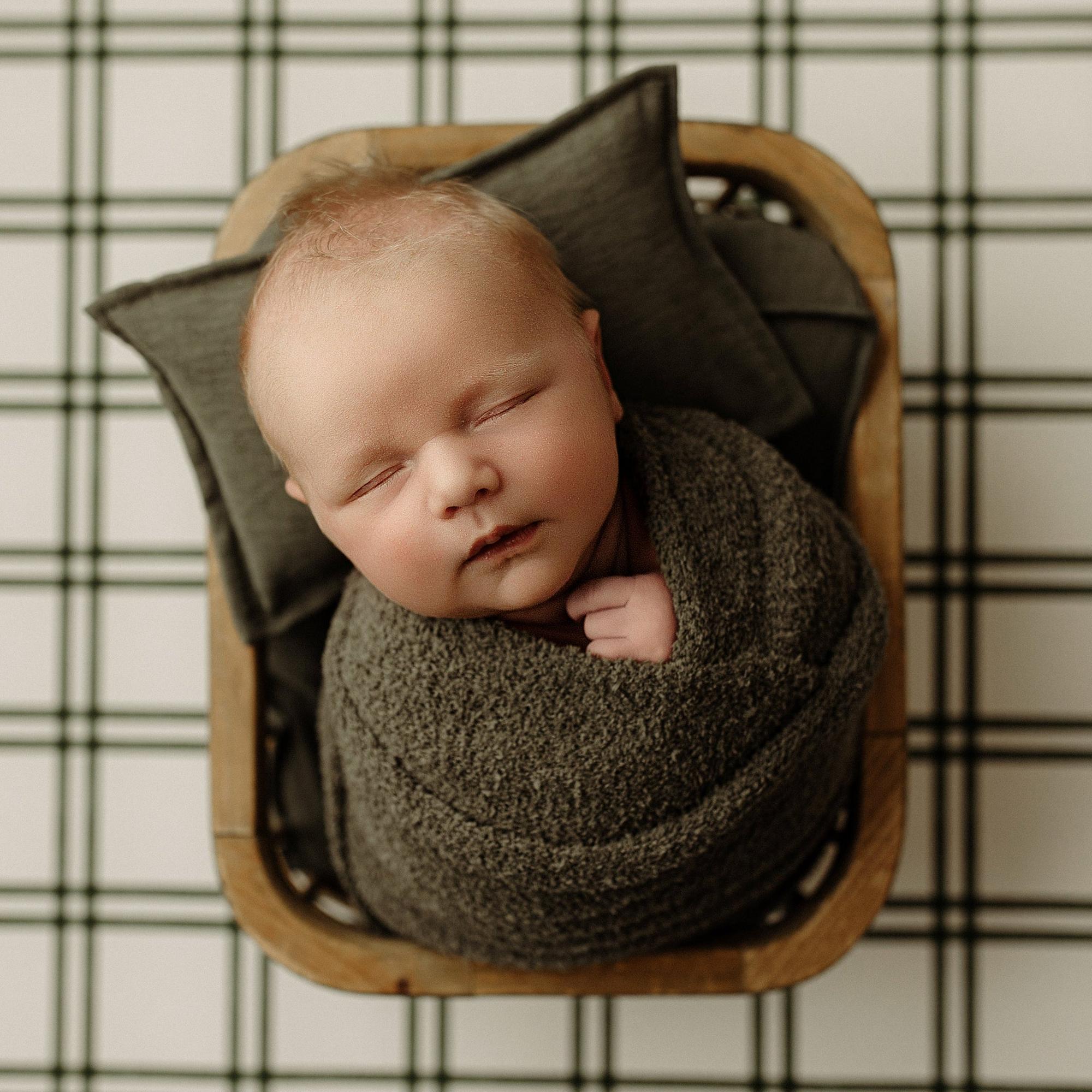 A sleeping newborn baby swaddled in a textured dark gray blanket, resting inside a wooden basket lined with dark gray pillows, set against a black and white plaid blanket background.