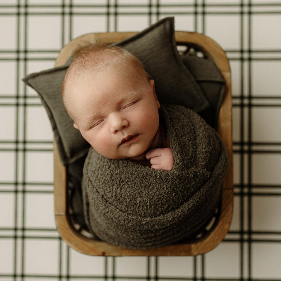 A sleeping newborn baby swaddled in a textured dark gray blanket, resting inside a wooden basket lined with dark gray pillows, set against a black and white plaid blanket background.