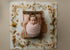 Newborn wrapped in a soft, strawberry milk pink open-knit tatter wrap with a matching floral tieback on a rustic wooden bed.