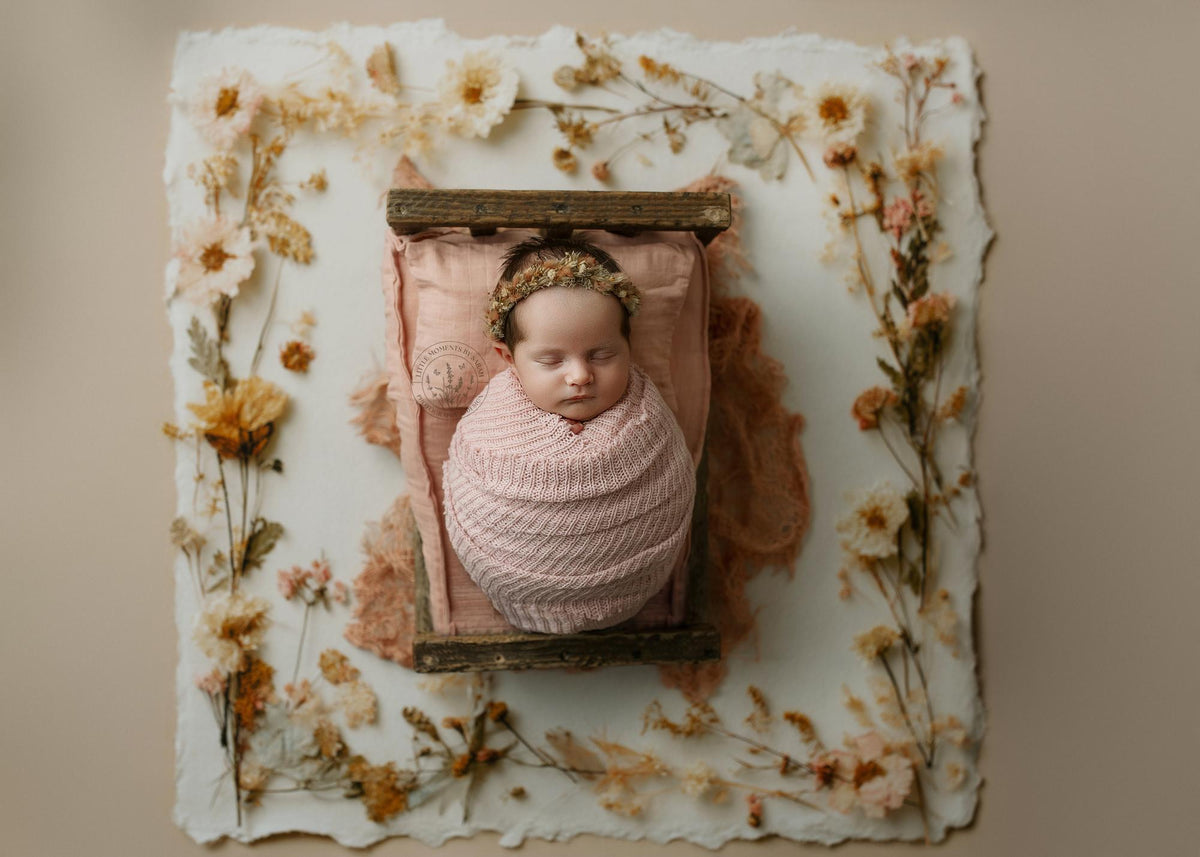 Newborn wrapped in a soft, strawberry milk pink open-knit tatter wrap with a matching floral tieback on a rustic wooden bed.