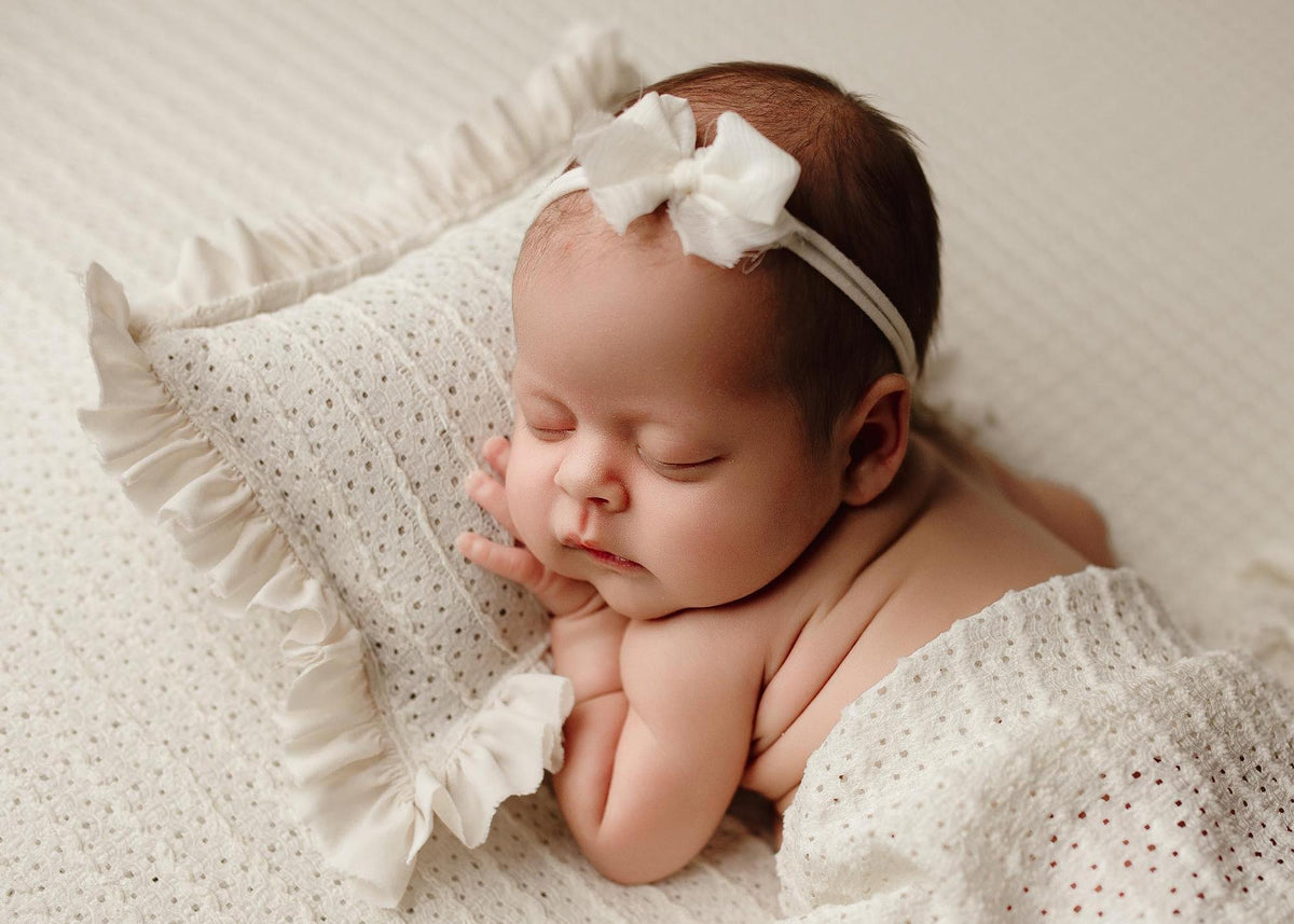 Newborn sleeping on a textured off-white Veda posing fabric and matching ruffled pillow with a delicate bow headband.