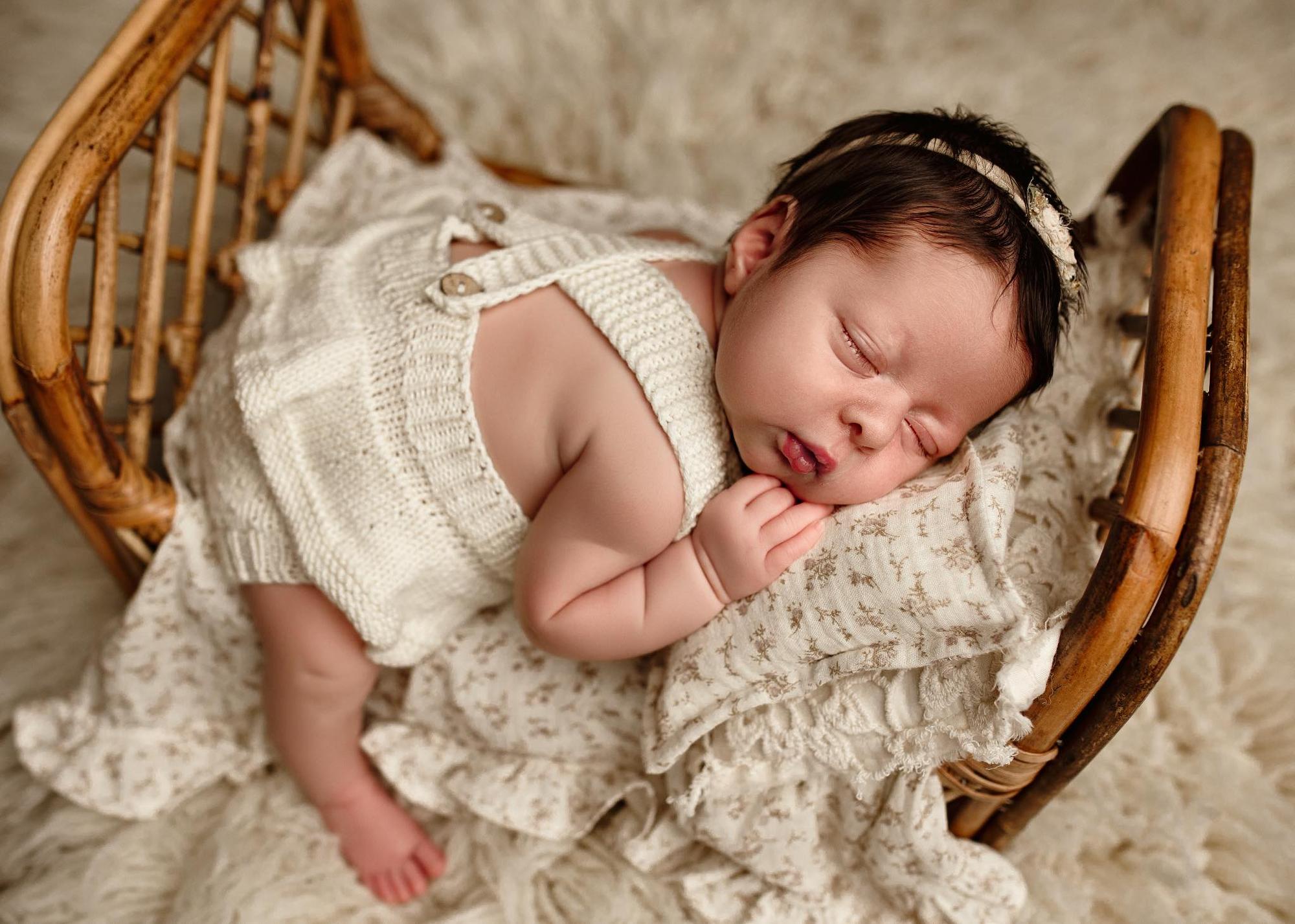 Newborn sleeping on a floral sheet and pillowcase set with ruffles inside a small rattan bed prop.