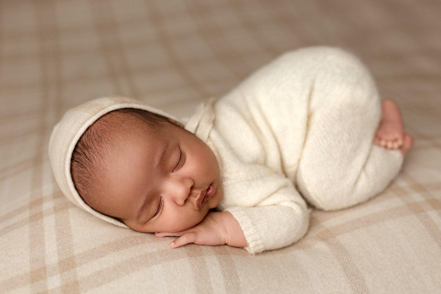 Newborn wearing a soft, cream-colored knit bonnet and matching romper, sleeping on a neutral plaid photography backdrop.