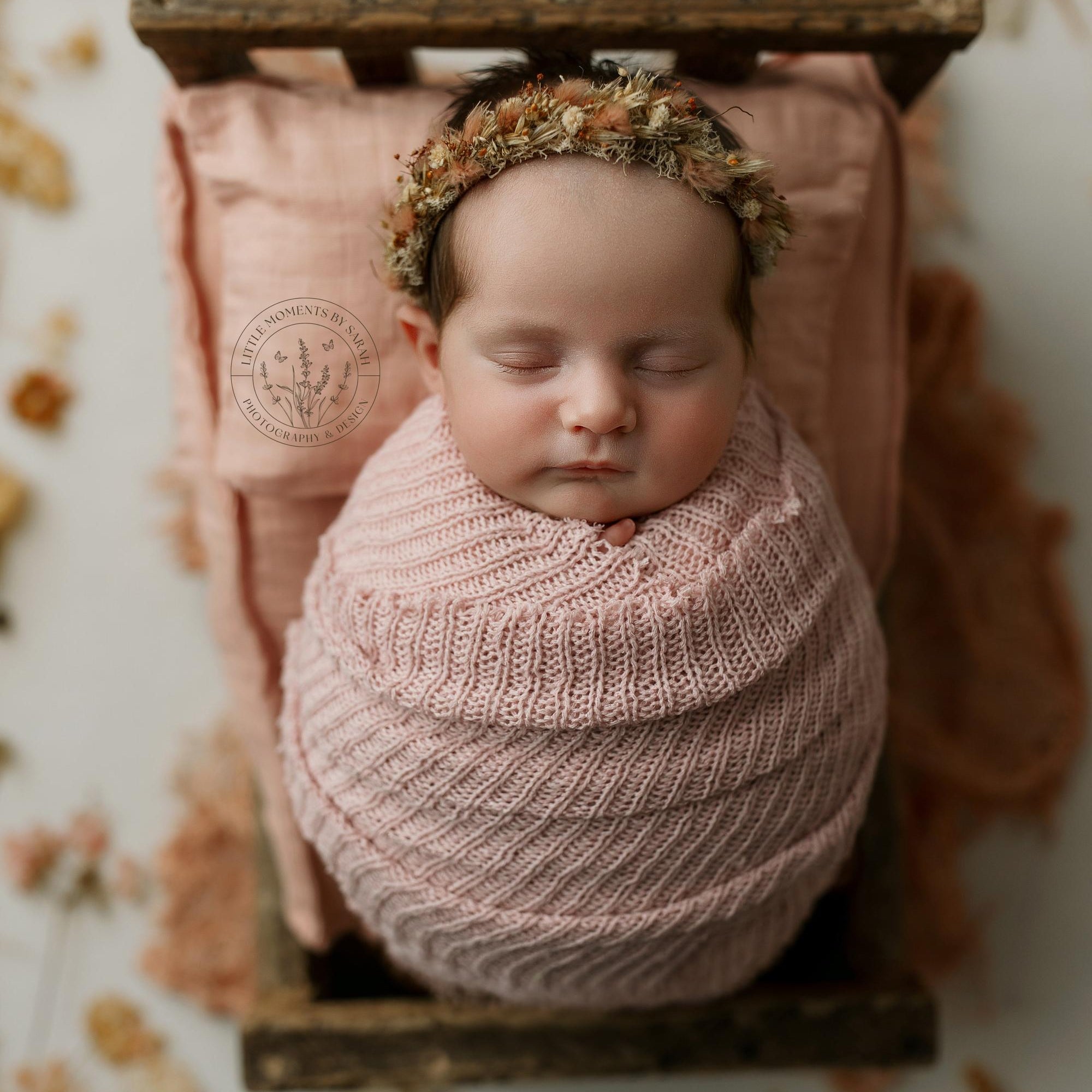 Newborn wrapped in a soft, strawberry milk pink open-knit tatter wrap with a matching floral tieback in a wooden bed.