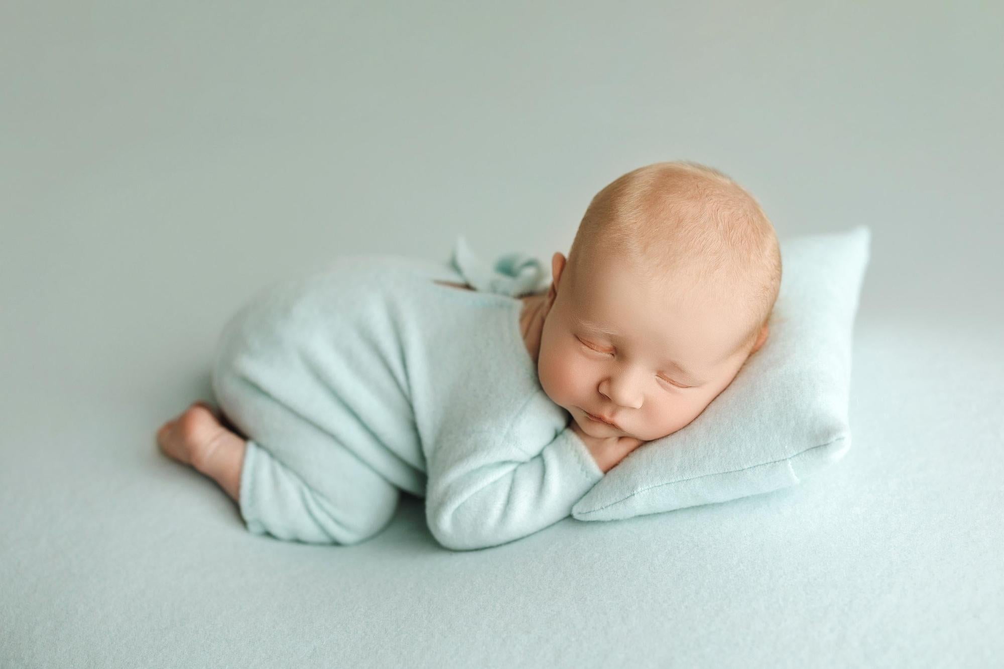 Newborn sleeping on a small pillow, wearing a soft, stretchy light blue romper from the Perry Collection.