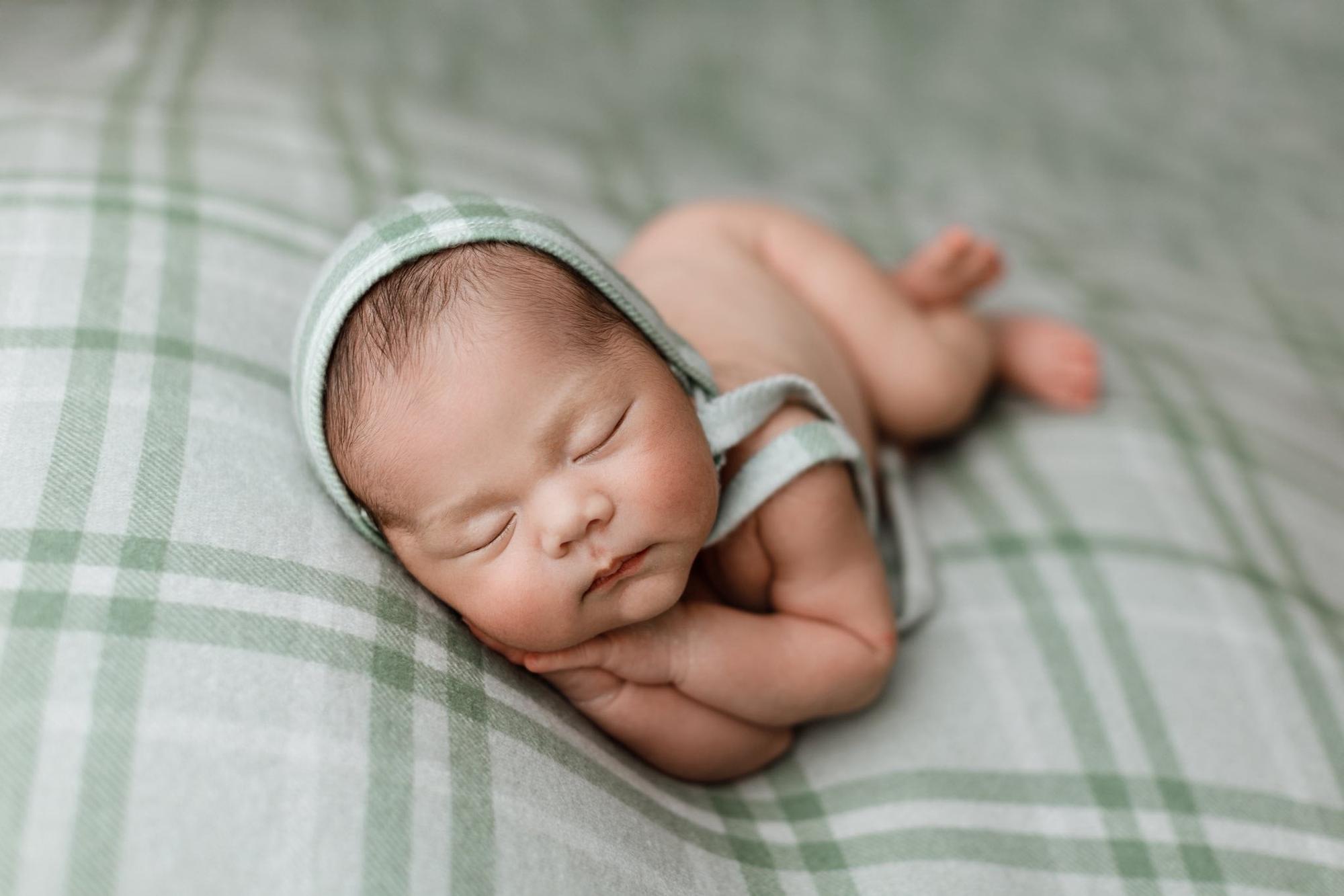Newborn sleeping on sage green and cream plaid fabric, wearing a matching vintage-style bonnet photography prop.