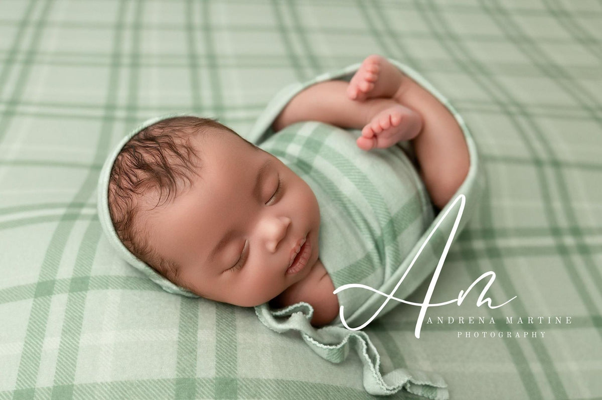 Newborn baby swaddled in a soft sage green and cream plaid fabric wrap for photography posing.