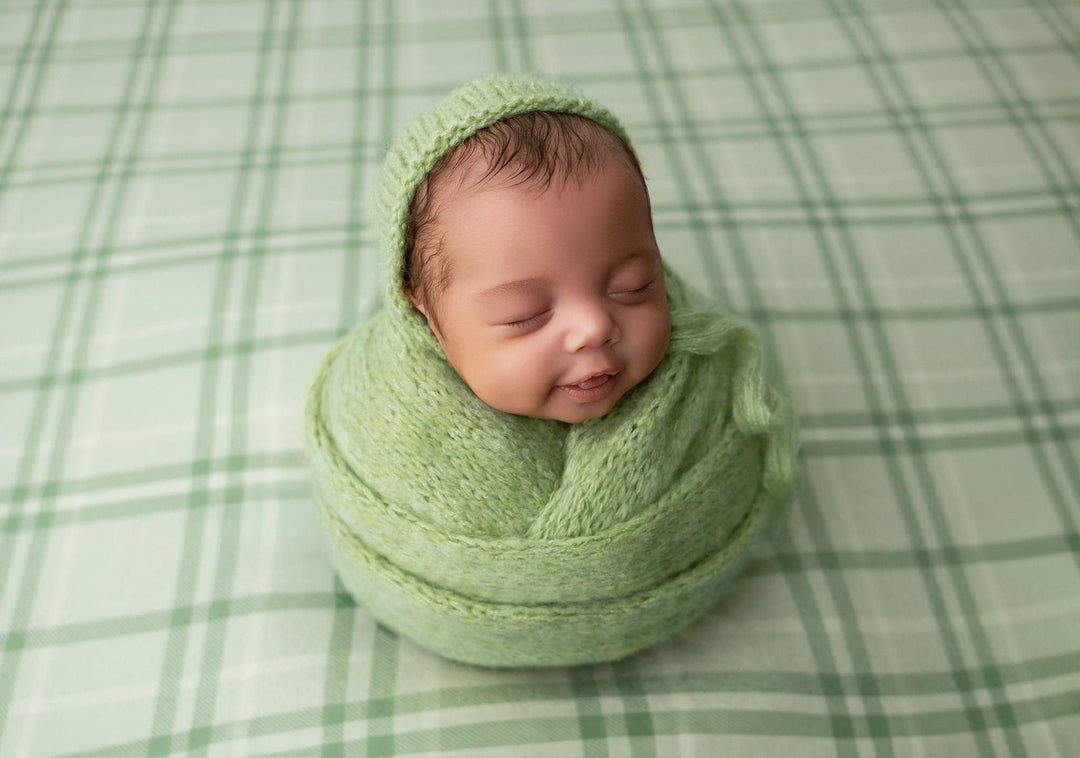 Newborn wrapped in a sage green knit bonnet and wrap, resting on a matching sage plaid fabric backdrop.