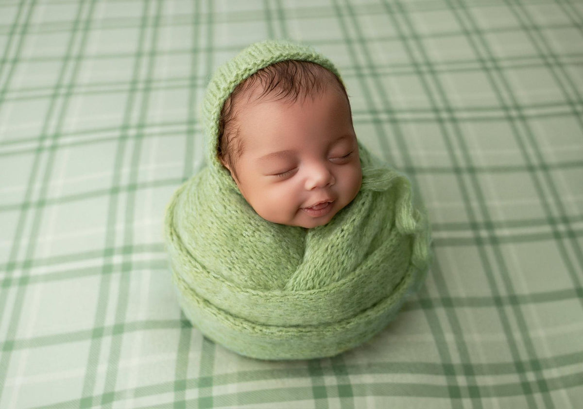 Newborn wrapped in a sage green knit bonnet and wrap, resting on a matching sage plaid fabric backdrop.