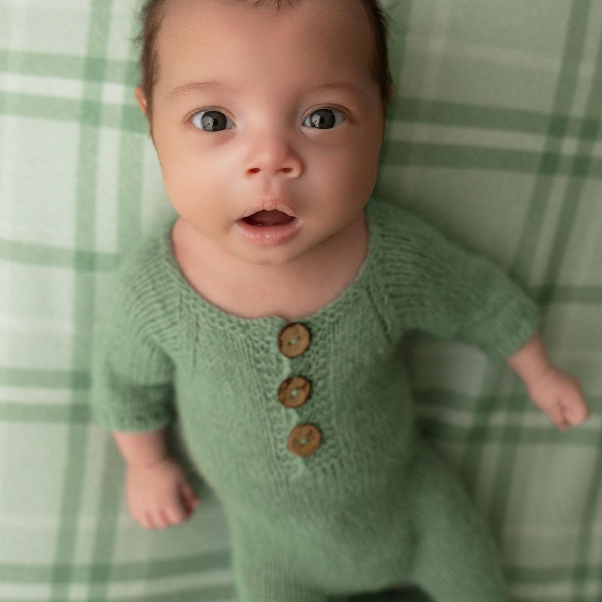 Newborn wearing a sage green knit romper on a matching sage and cream plaid fabric backdrop for newborn photography.