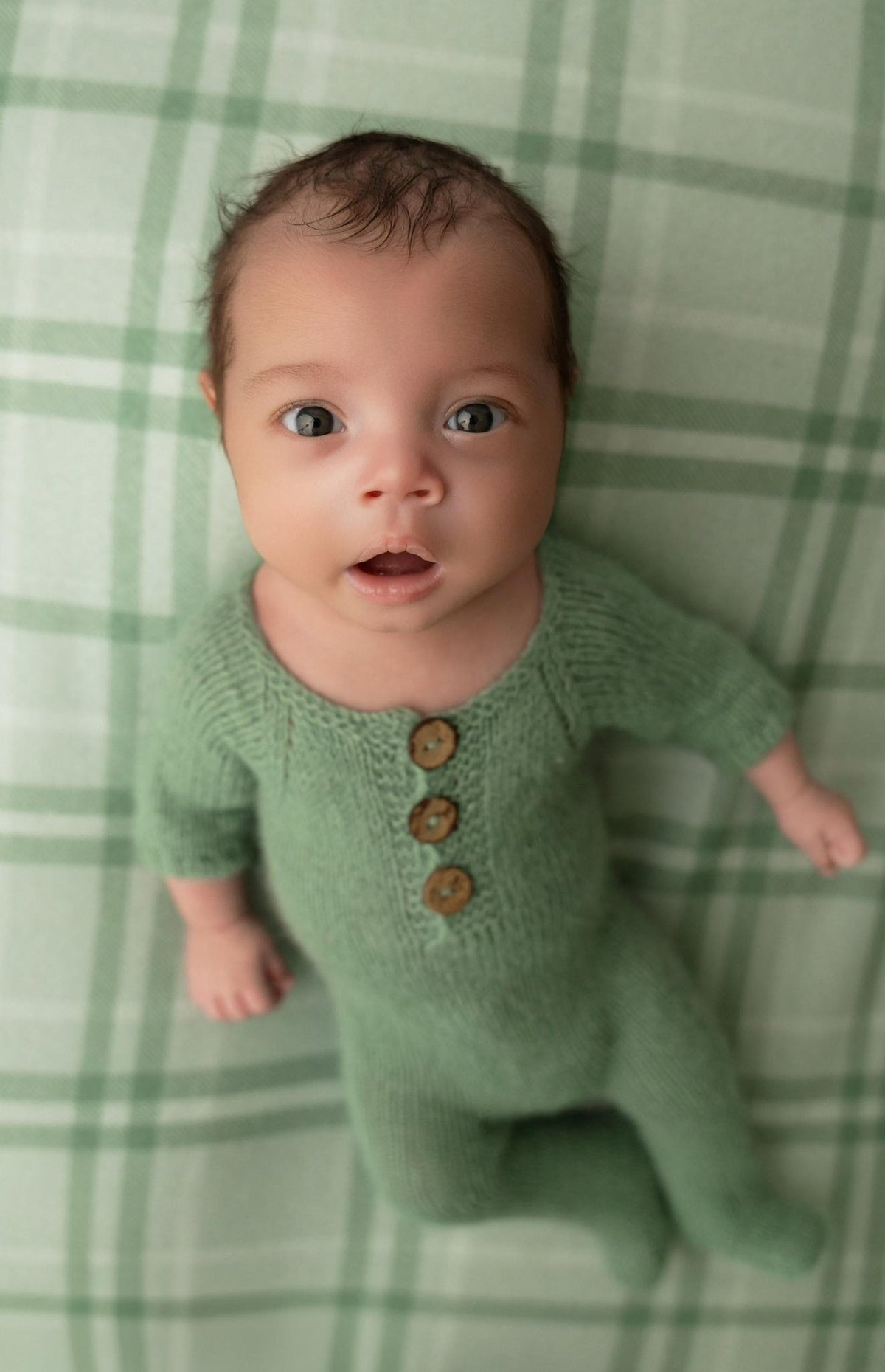 Newborn wearing a sage green knit romper on a matching sage and cream plaid fabric backdrop for newborn photography.