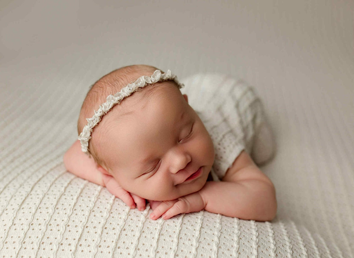 Newborn sleeping on a textured, off-white Veda knit posing fabric with a matching wrap and delicate lace headband.