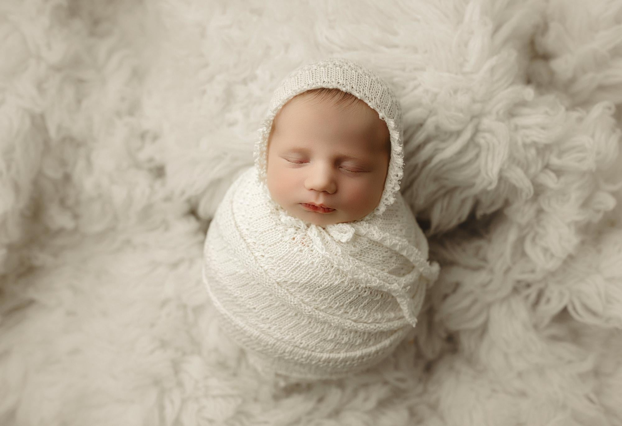 Newborn baby swaddled in an ivory open-knit wrap and matching bonnet, resting on a soft, textured photography backdrop.