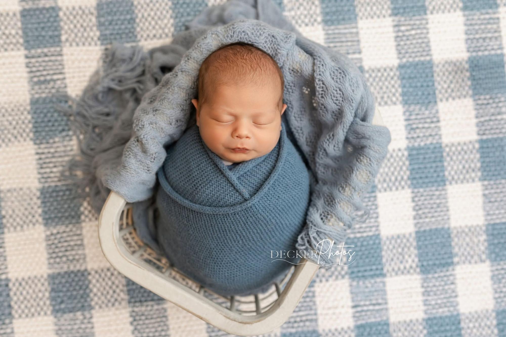 A sleeping newborn baby wrapped in a dusty blue knit swaddle and resting in a white basket lined with a matching blue knit blanket, set on the Willow Check poly paper backdrop by Little Moon Boutique.