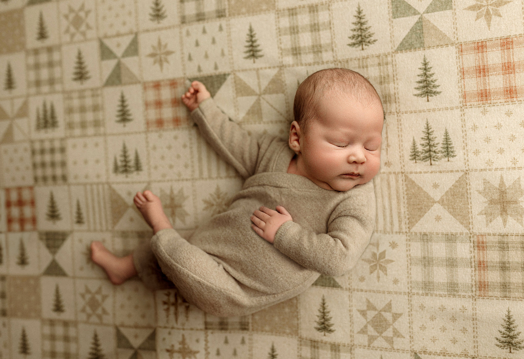 A sleeping newborn baby wearing a light taupe knit one-piece outfit, lying on a patterned quilt with a winter forest theme.