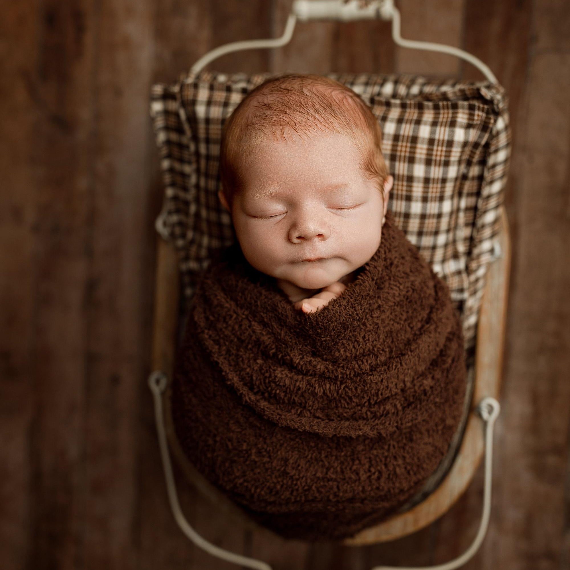 Newborn baby swaddled in a soft, fuzzy cocoa brown wrap, resting in a rustic prop basket with a plaid pillow.