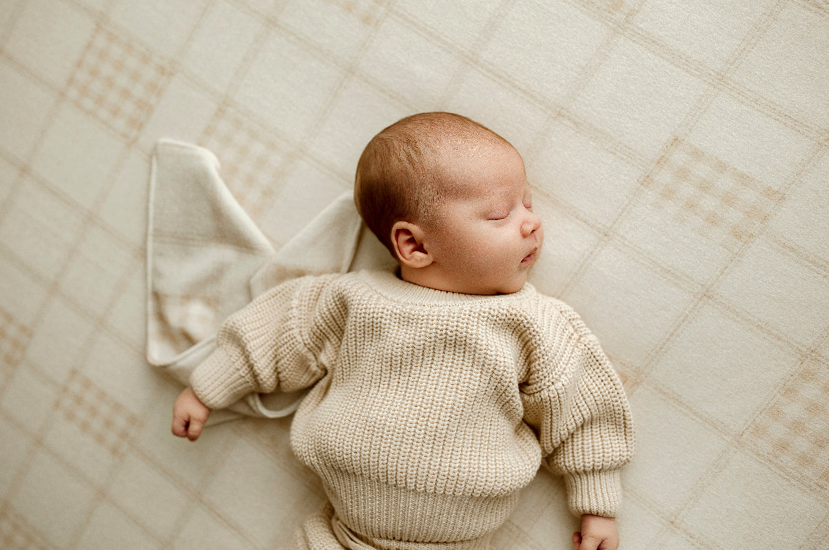 Close-up of sleeping newborn wearing a textured oatmeal knit sweater on Grandpa's Twin beige plaid posing fabric with a coordinating mini blankie.