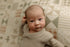 A close-up of a newborn baby lying on a patterned quilted blanket, wearing a soft beige knit romper.