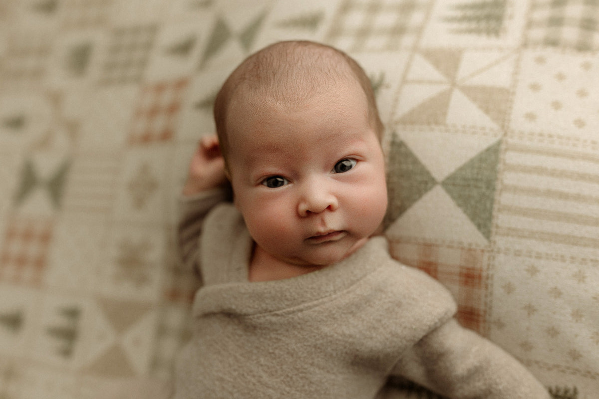A close-up of a newborn baby lying on a patterned quilted blanket, wearing a soft beige knit romper.