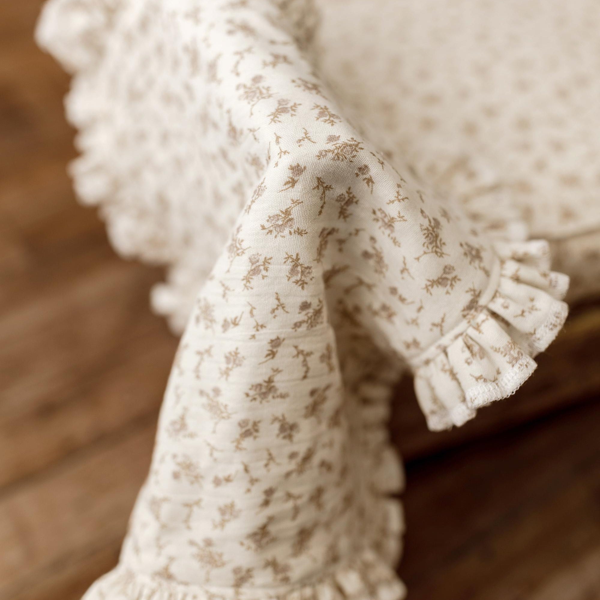 A close-up of a cream-colored fabric newborn photography mini blanket with a delicate beige floral pattern and ruffled edges, draped over a wooden surface.