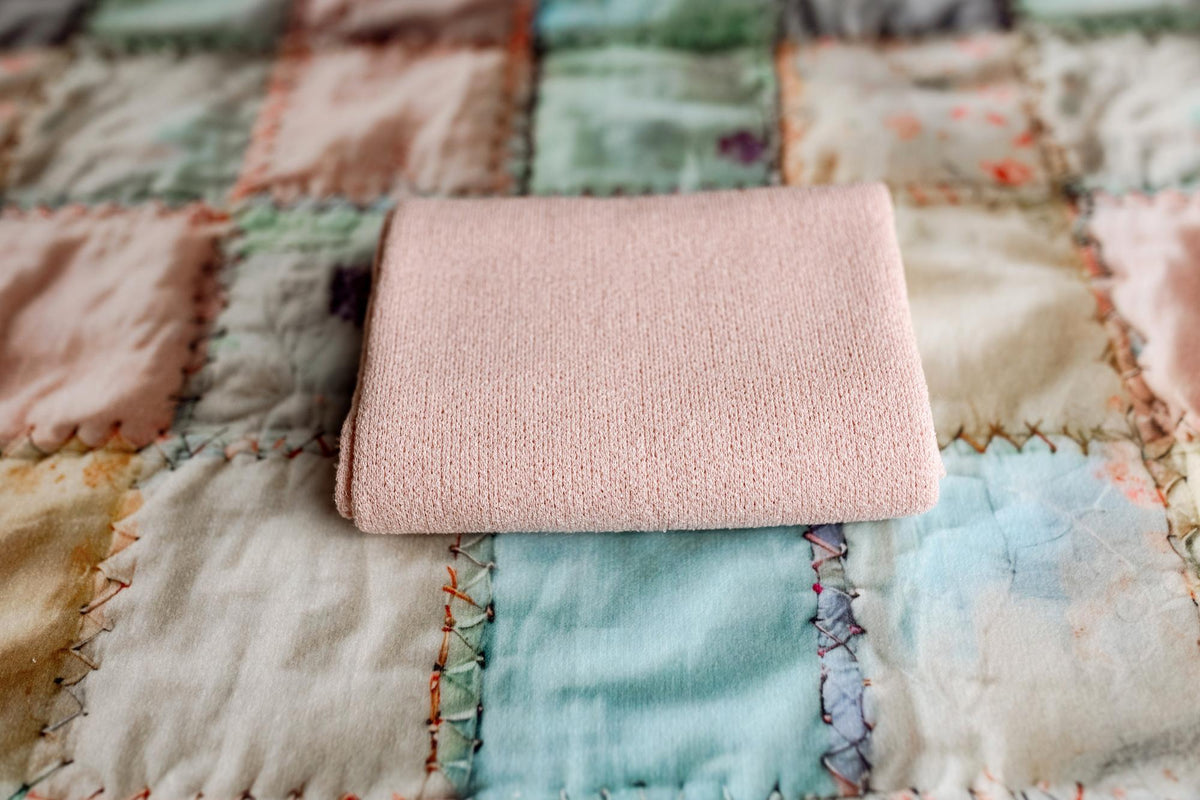 Folded carnation pink stretchy knit newborn posing wrap resting on a pastel patchwork quilt.