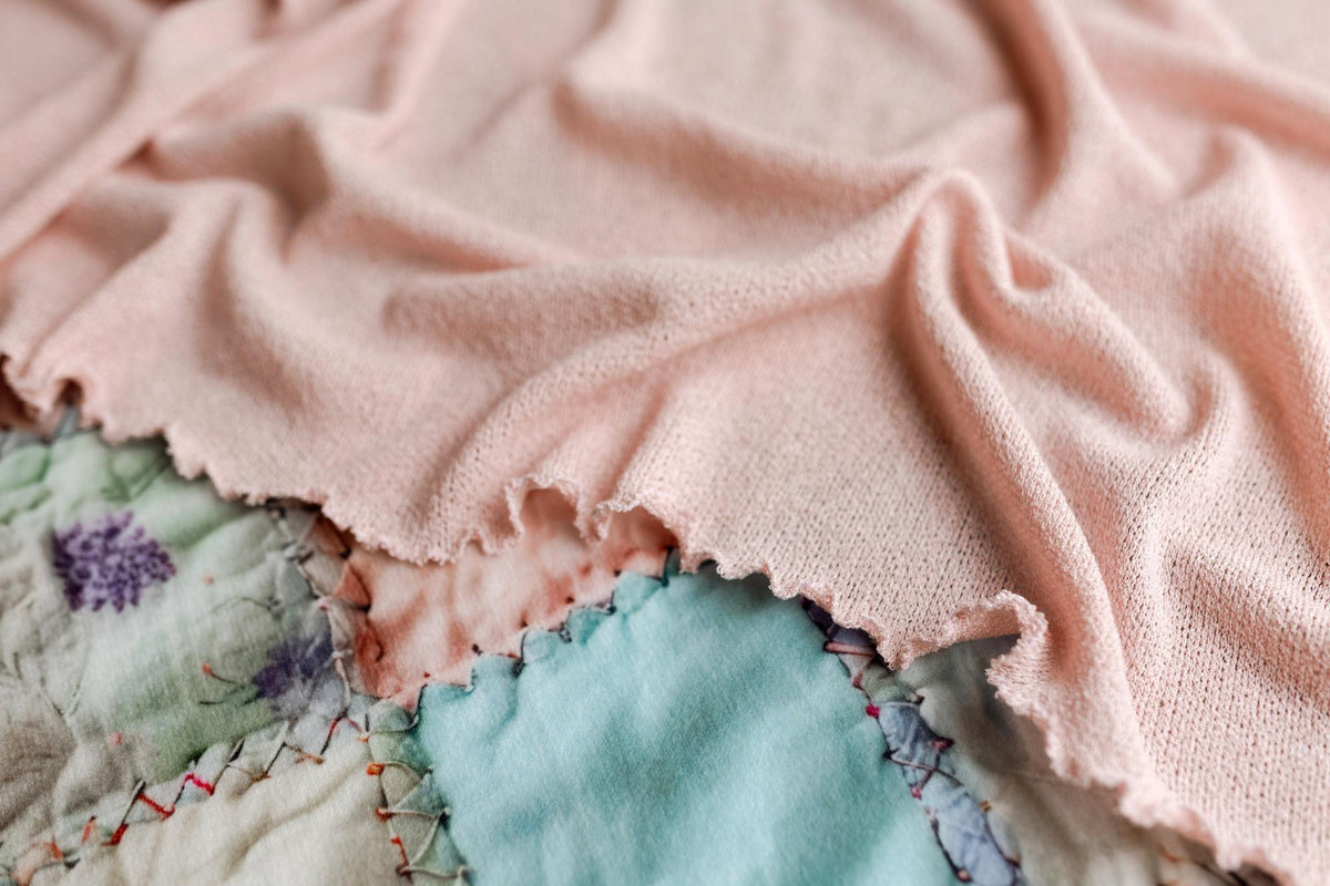 Soft carnation pink stretchy knit newborn photography wrap with ruffled edges draped over a pastel patchwork quilt.