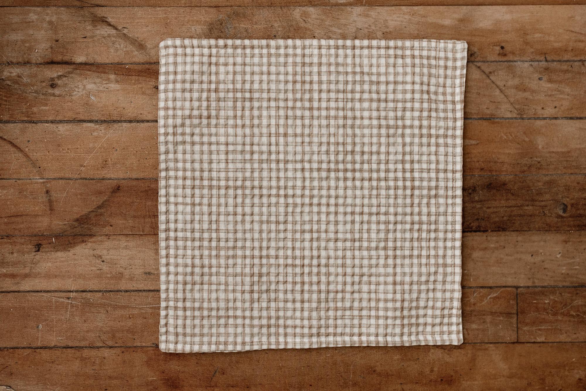 A square beige and brown gingham checkered newborn photography mini blanket laid flat on a rustic wooden plank surface.