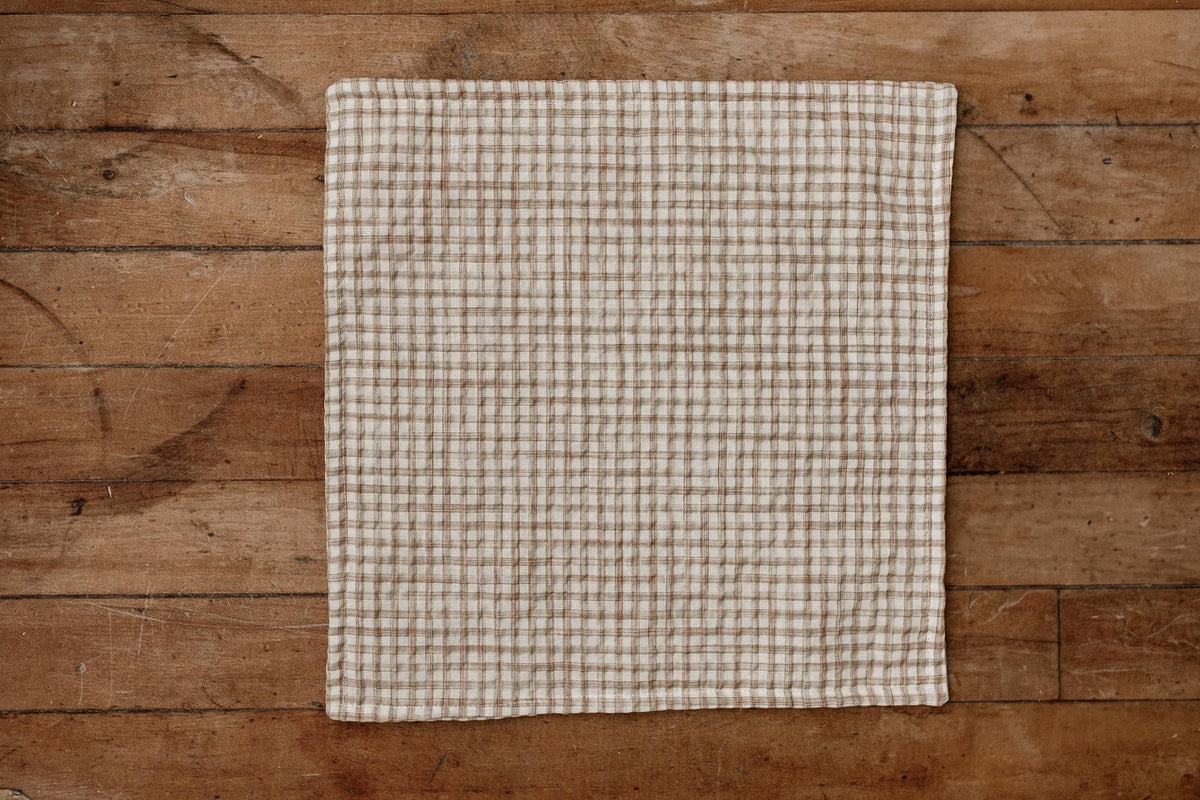 A square beige and brown gingham checkered newborn photography mini blanket laid flat on a rustic wooden plank surface.