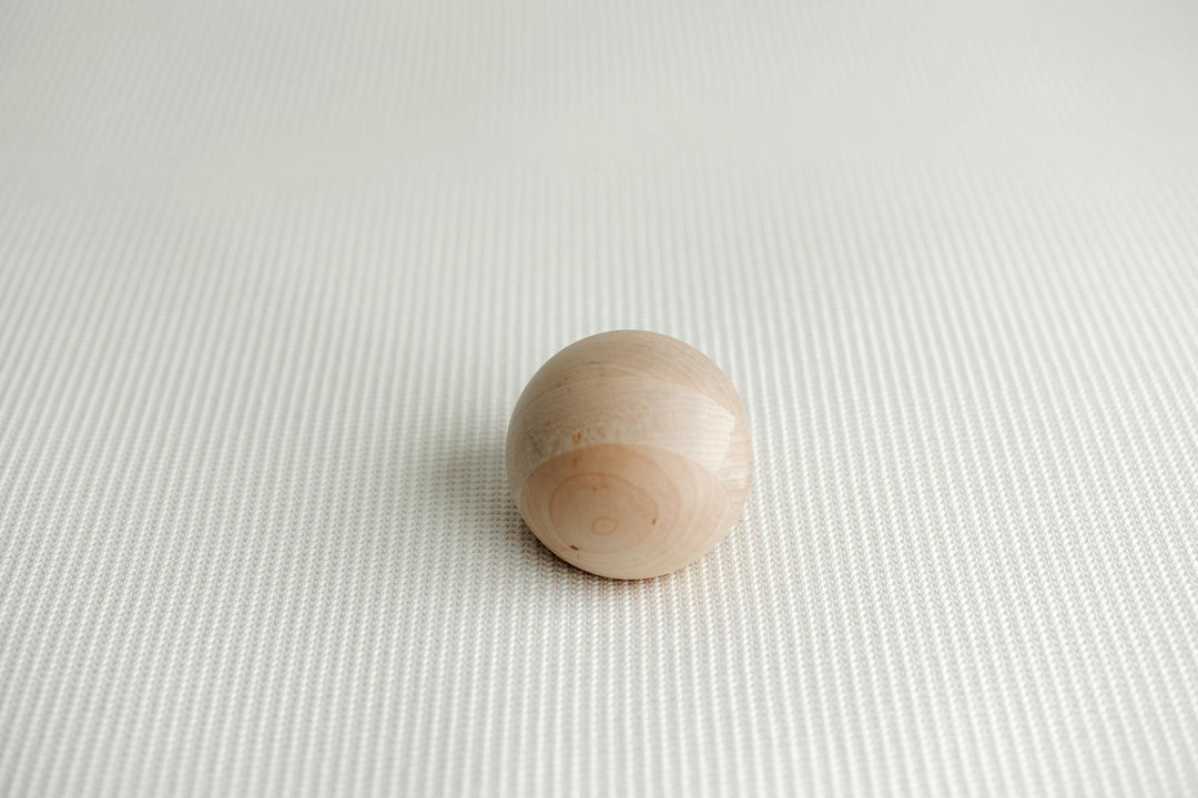 A small, smooth, spherical wooden bead or knob in a light natural wood finish, resting on a textured white newborn posing fabric.