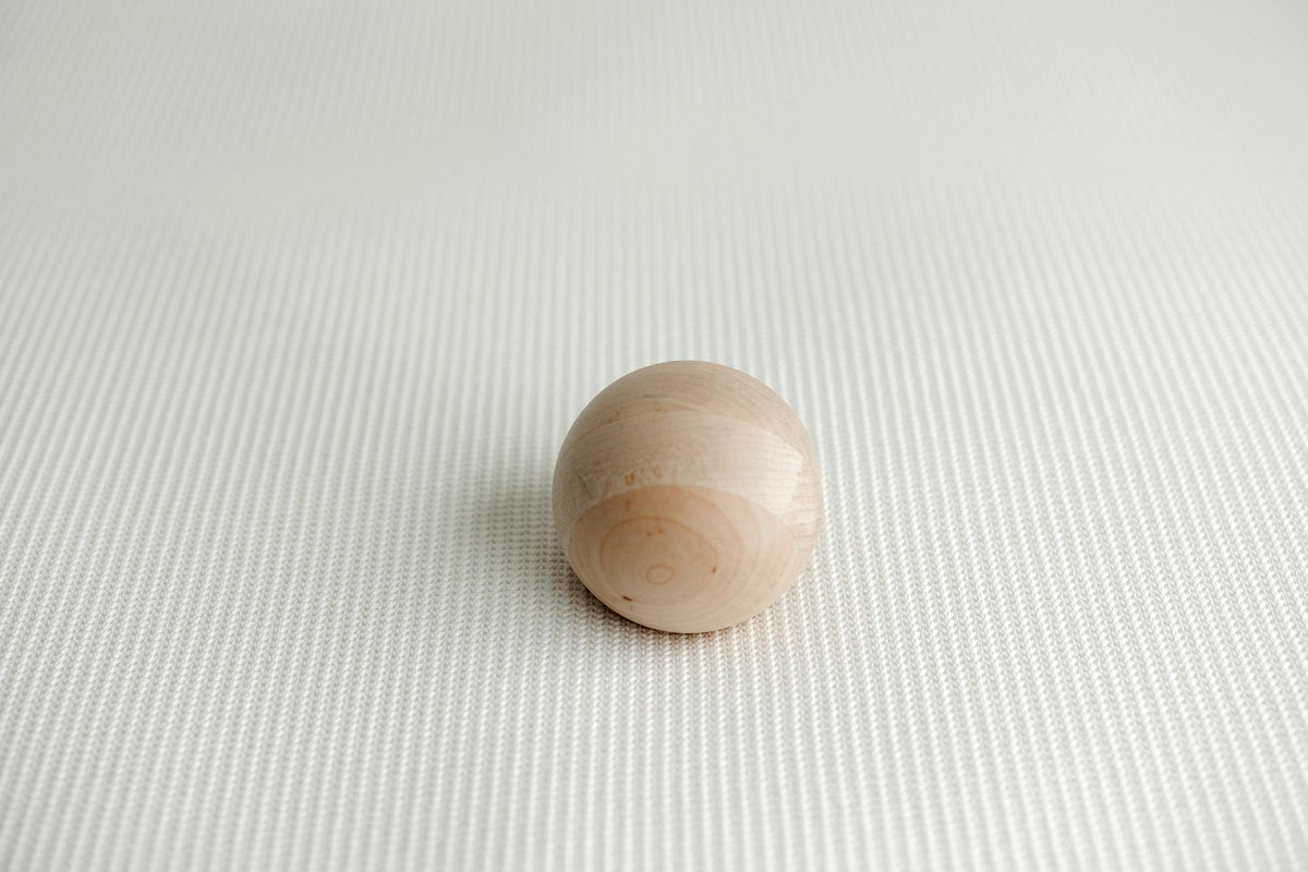 A small, smooth, spherical wooden bead or knob in a light natural wood finish, resting on a textured white newborn posing fabric.