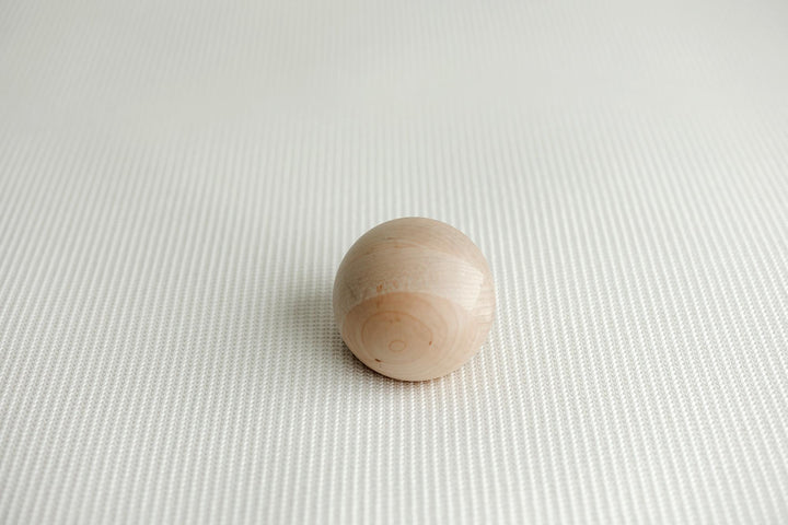 A small, smooth, spherical wooden bead or knob in a light natural wood finish, resting on a textured white newborn posing fabric.