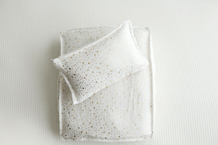 A white textured pillow with small gold speckles resting on top of a matching white quilted blanket with gold speckles, set against a light gray ribbed background.