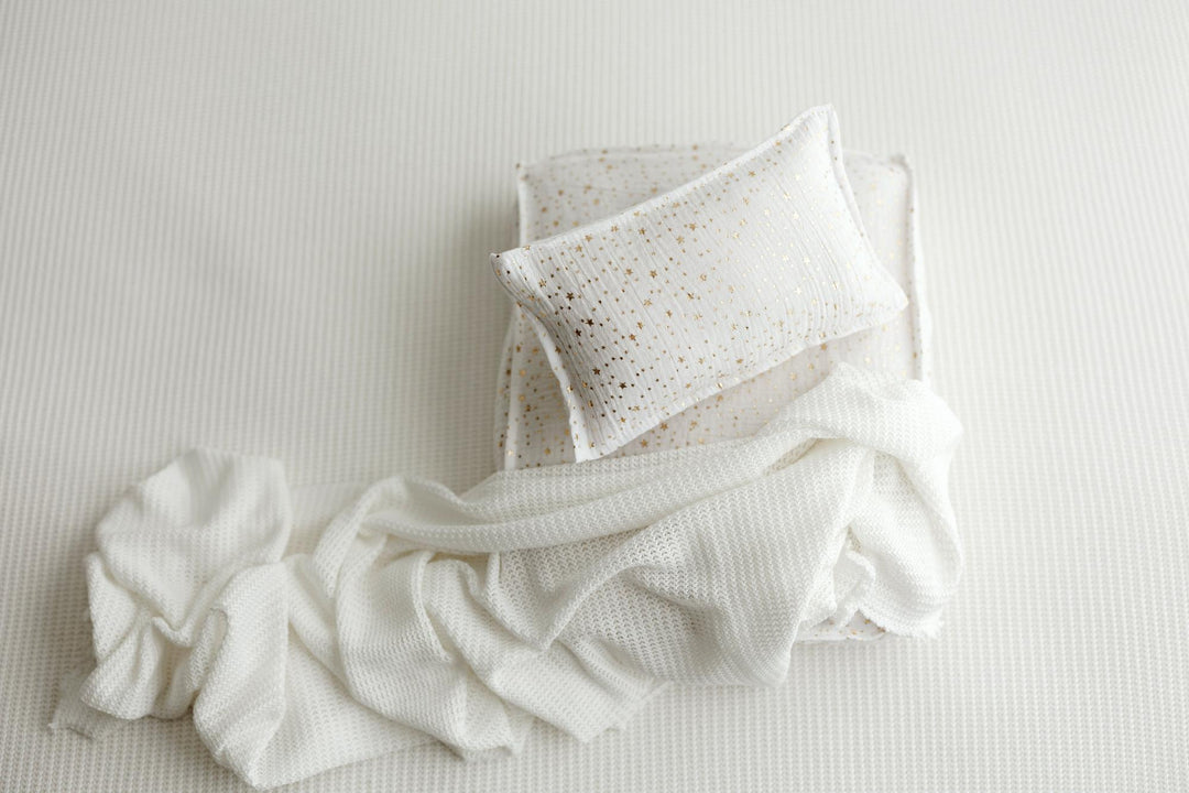 A white textured pillow with small gold speckles resting on top of a matching white waffle-knit blanket, arranged on a white ribbed fabric background.