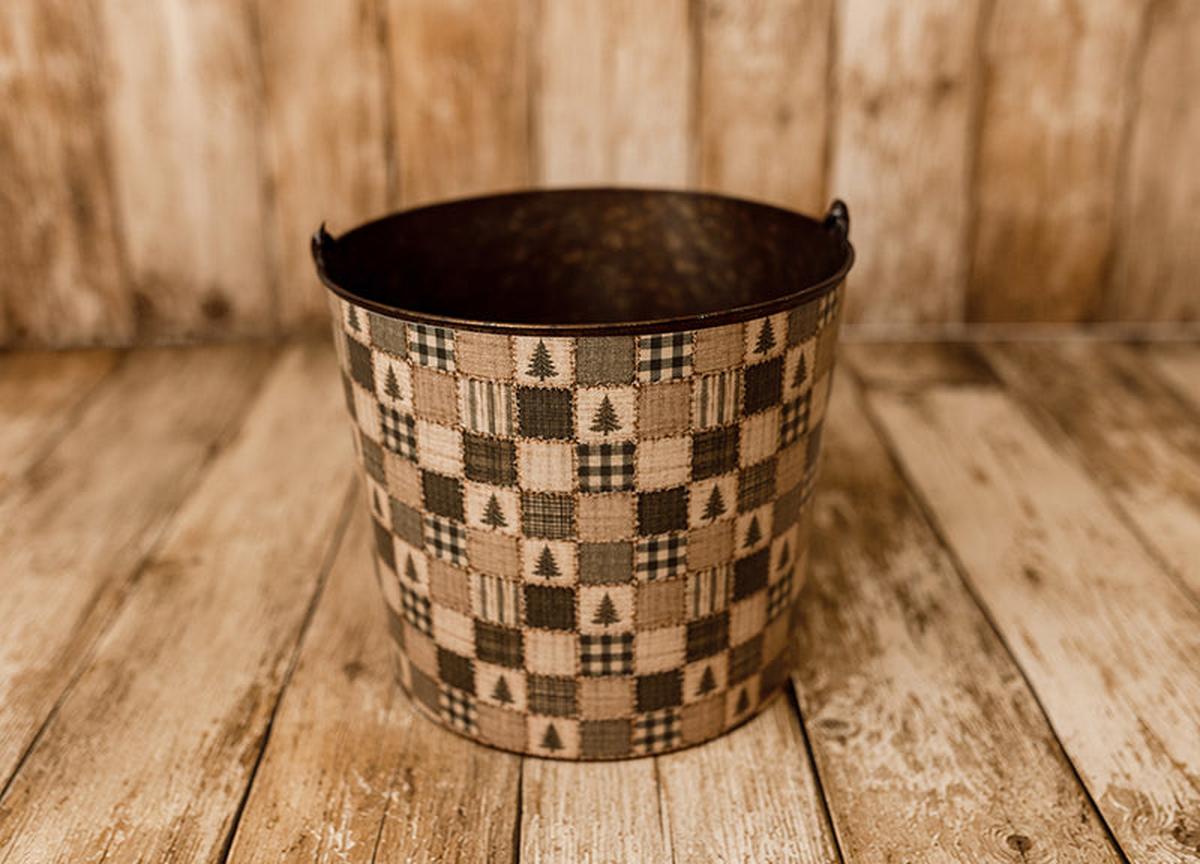 Tree Farm Christmas newborn photography bucket cover featuring vintage patchwork squares with evergreen trees, black and cream buffalo plaid, ticking stripes, and rustic neutral tones, displayed on a metal posing bucket.