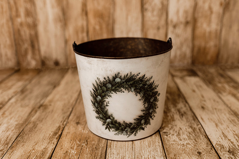December Morning Christmas newborn photography bucket cover featuring a rustic evergreen and eucalyptus wreath on a softly distressed cream background, displayed on a vintage metal posing bucket against a wooden farmhouse backdrop.